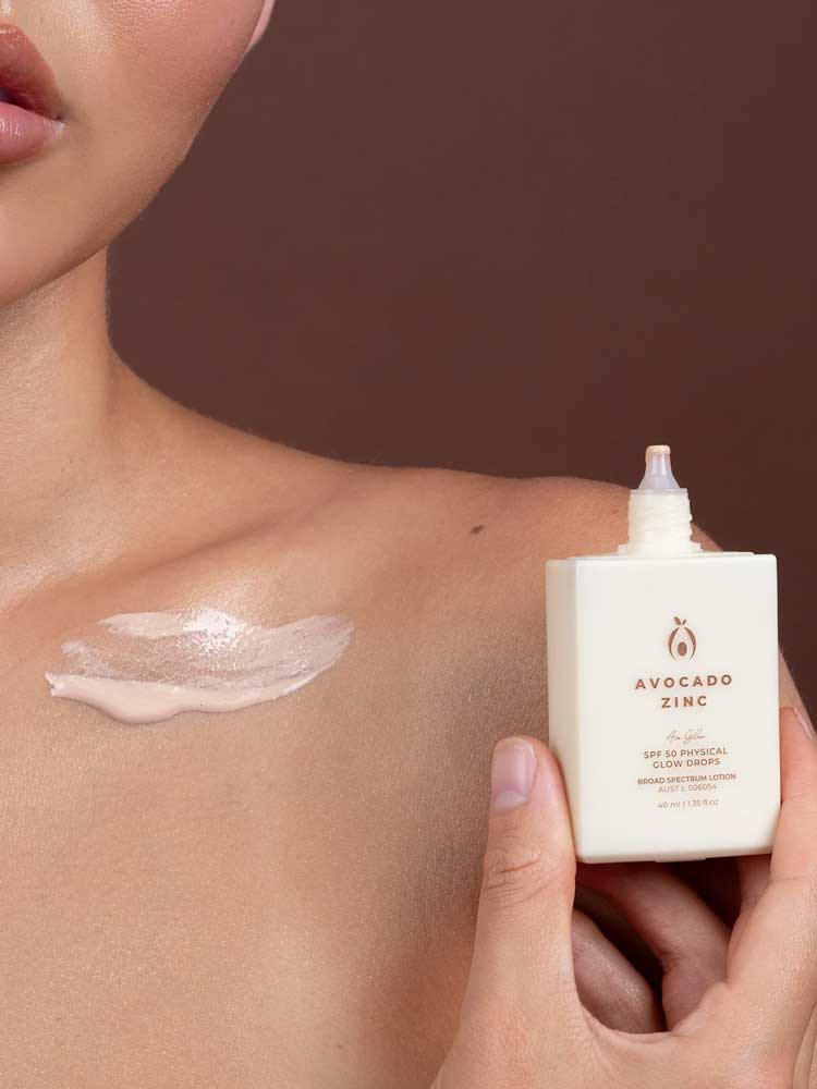 A person holds a bottle of Avocado Zinc SPF50 Physical Glow Drops 40ml near their collarbone, displaying a dab of the luminous formula on their shoulder against a brown background.