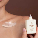 A person holds a bottle of Avocado Zinc SPF50 Physical Glow Drops 40ml near their collarbone, displaying a dab of the luminous formula on their shoulder against a brown background.
