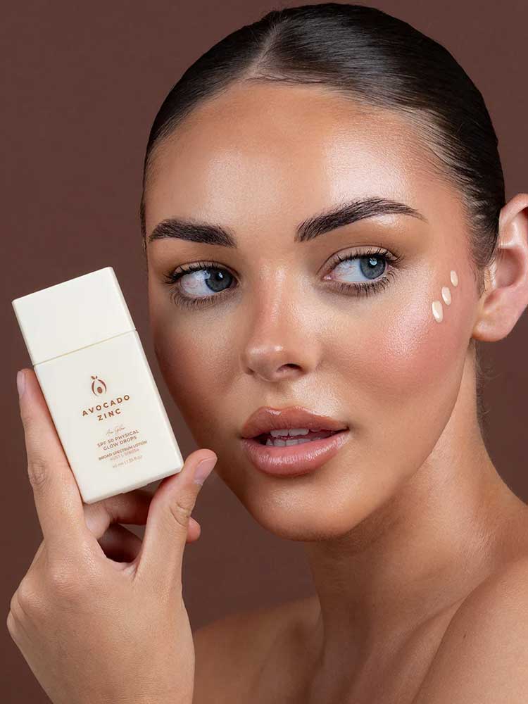 A woman with clear, glowing skin holds a bottle of Avocado Zinc SPF50 Physical Glow Drops 40ml near her face, with three dots of cream on her cheek, set against a brown background.