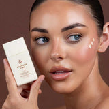 A woman with clear, glowing skin holds a bottle of Avocado Zinc SPF50 Physical Glow Drops 40ml near her face, with three dots of cream on her cheek, set against a brown background.