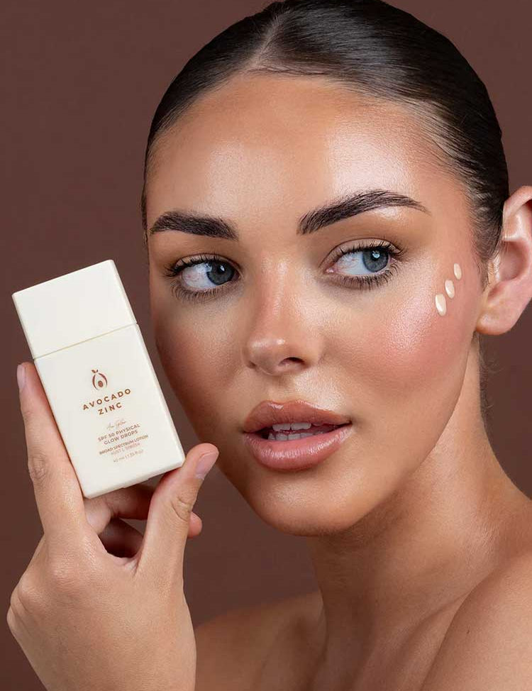 A woman with clear, glowing skin holds a bottle of Avocado Zinc SPF50 Physical Glow Drops 40ml near her face, with three dots of cream on her cheek, set against a brown background.