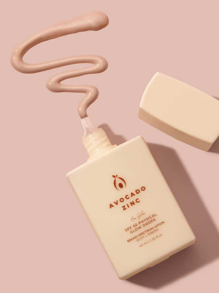 A bottle of Avocado Zinc SPF50 Physical Glow Drops 40ml in beige sits on a pink surface with the cap off, and the creamy product is dispensed in a wavy line above it.