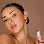 Model wearing Avocado Zinc SPF50 Natural Lip & Cheek Tint in Nude and holding the tint beside her face.