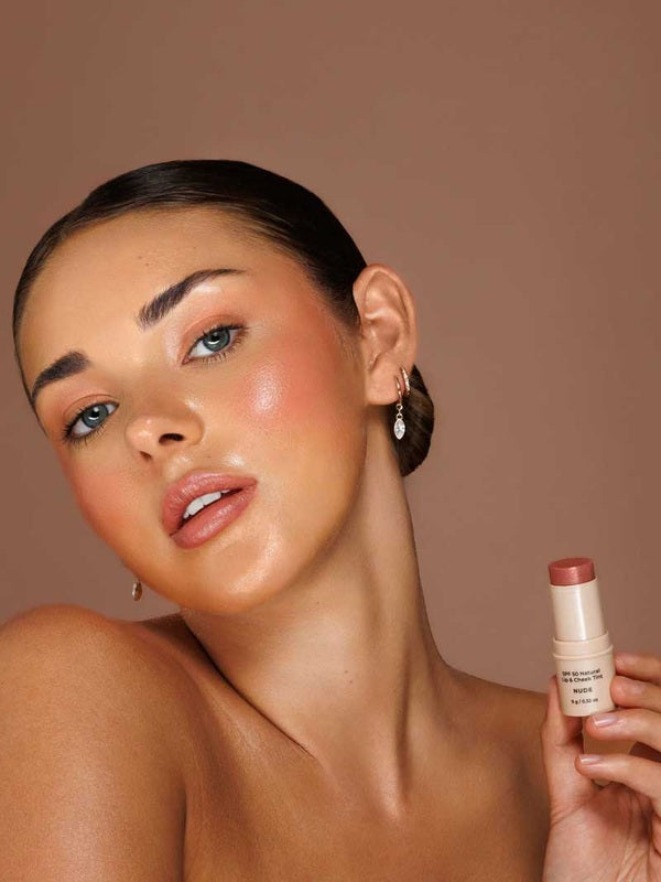 Model wearing Avocado Zinc SPF50 Natural Lip & Cheek Tint in Nude and holding the tint beside her face.