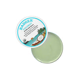 An open tin of Badger Coconut After Sun Balm 56g displays light green balm. The lid features product details and a tropical scene with coconut, palm trees, and water, highlighting soothing relief for sunburned skin.