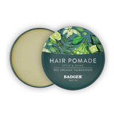 A round tin of Badger Hair Pomade 56g, featuring green botanical designs and labeled "Hair Pomade, Style & Shine, 85% Organic Ingredients," sits with its lid partially open.