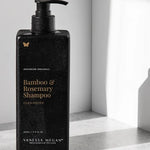 Black rectangular Vanessa Megan Bamboo & Rosemary Shampoo bottle with a black pump and gold lettering, covered in droplets on a sunlit white ledge.