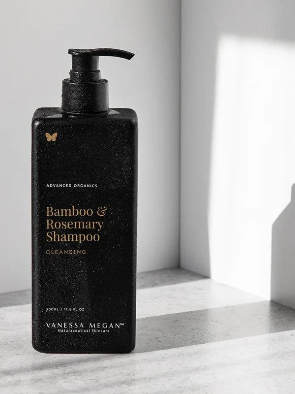 Black rectangular Vanessa Megan Bamboo & Rosemary Shampoo bottle with a black pump and gold lettering, covered in droplets on a sunlit white ledge.