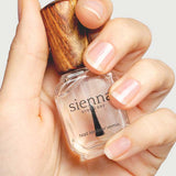 A hand displays a bottle of Sienna Byron Bay Nail Polish - Base Coat with a wooden cap. The neatly manicured natural nails are ready for the clear, glossy base coat formula.
