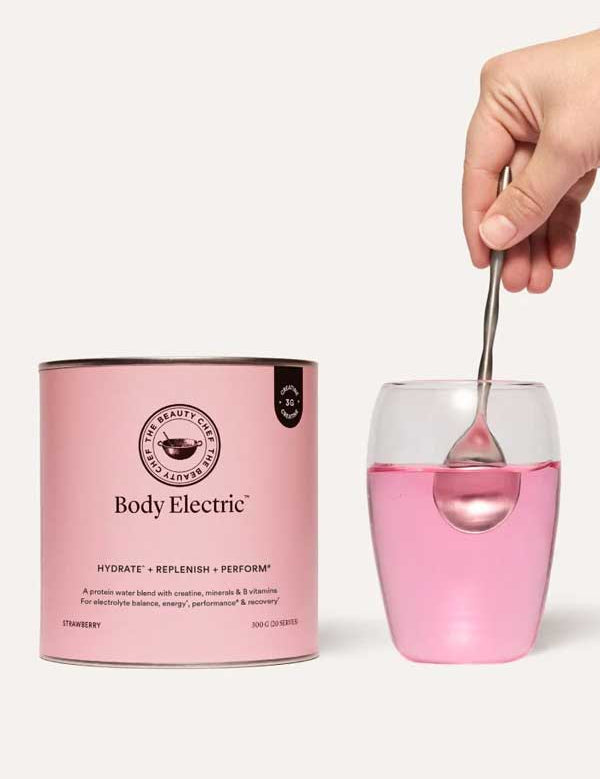 A hand stirs a spoon in a glass of pink drink beside The Beauty Chef Body Electric™ Protein Water + Creatine 320g canister, featuring plant protein, electrolytes, and a tasty strawberry flavor.