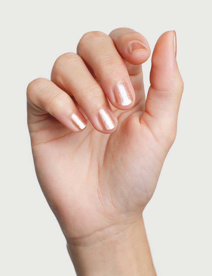A close-up of a hand with fingers slightly curled, featuring nails painted in Sienna Byron Bay Nail Polish - Believe, a shiny light rose peach shade, set against a plain light background.