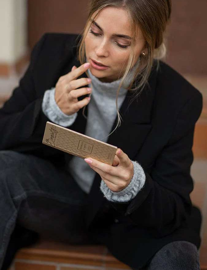 A blonde woman in a loose bun sits on steps, holding a Benecos Beauty ID Palette Lip & Cheek Refill - Aperitif Kiss 3g and examining its vegan packaging. She wears a black blazer with a grey sweater, touching her lips thoughtfully.