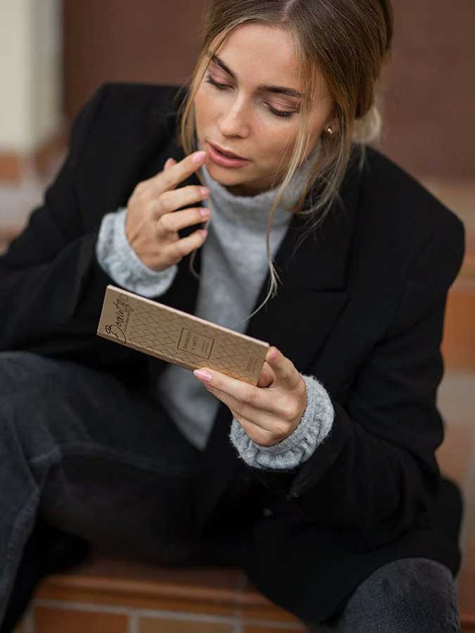 A blonde woman in a loose bun sits on steps, holding a Benecos Beauty ID Palette Lip & Cheek Refill - Aperitif Kiss 3g and examining its vegan packaging. She wears a black blazer with a grey sweater, touching her lips thoughtfully.