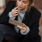 A woman in a black blazer and gray sweater sits while applying Benecos Beauty ID Palette Lip & Cheek Refill – Velvet Rose 3g, thoughtfully holding its rectangular box.