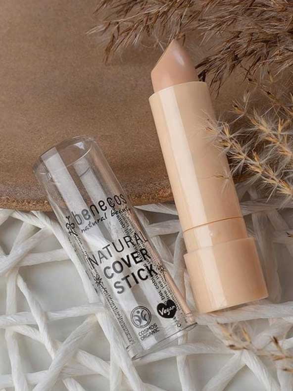 A beige Benecos Cover Stick 4.5g with its clear cap off sits on a woven white surface next to dried grasses. This high coverage concealer is vegan and certified organic by Benecos.