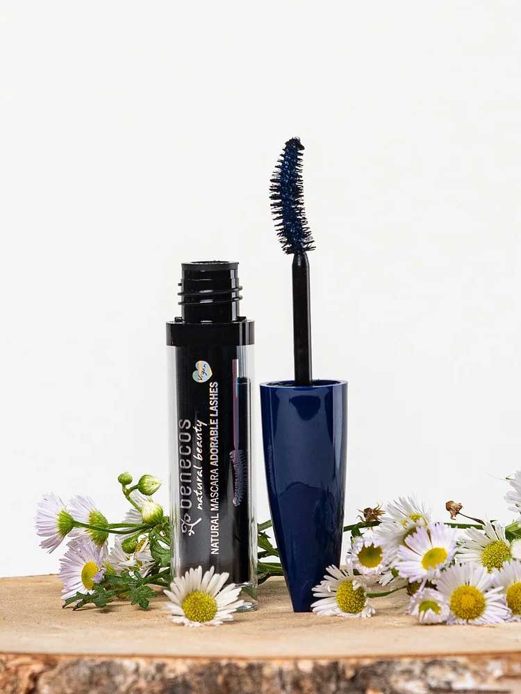 An open tube of Benecos Mascara Adorable Lashes Deep Ocean 5.5ml with a blue wand rests among white and yellow daisies on a light background, ideal for naturally lengthening lashes.