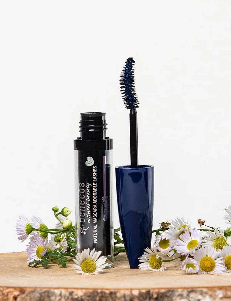 An open tube of Benecos Mascara Adorable Lashes Deep Ocean 5.5ml with a blue wand rests among white and yellow daisies on a light background, ideal for naturally lengthening lashes.