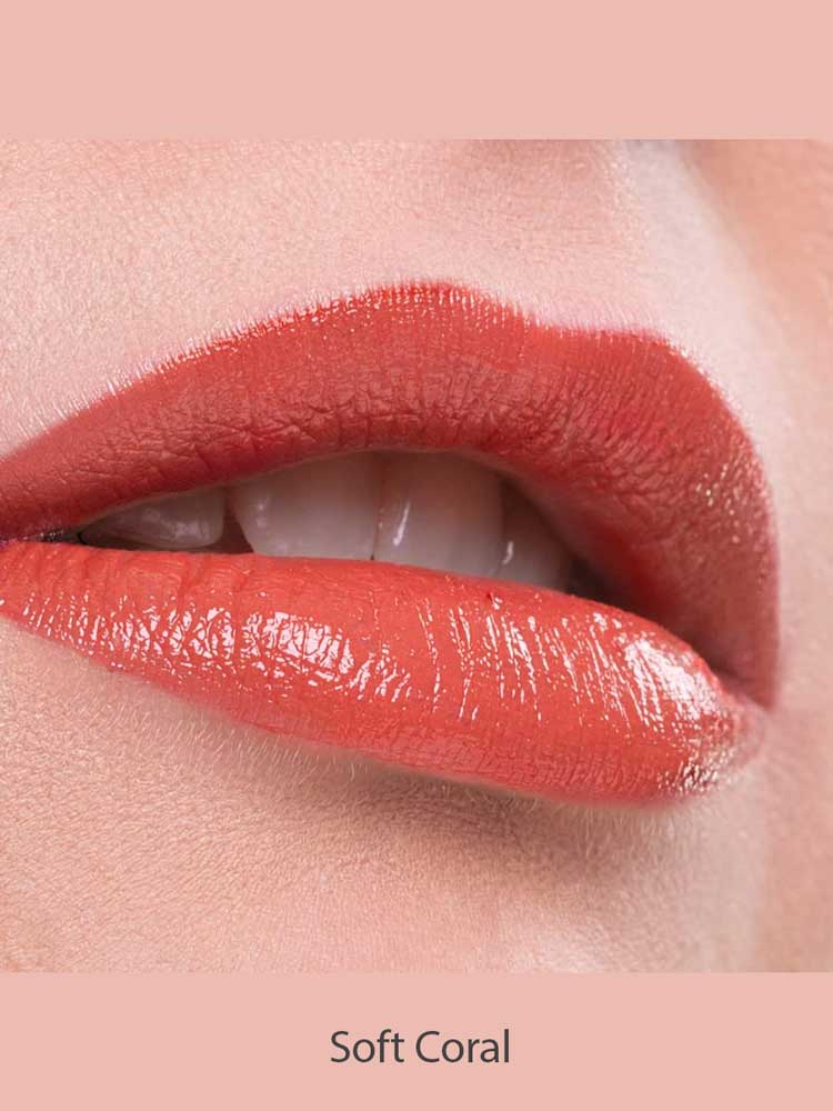 Close-up of lips wearing Benecos Natural Lipsticks 4.5g in a glossy, hydrating coral shade, slightly parted to reveal teeth. The background and area around the lips are a soft coral hue. Text below reads Soft Coral.