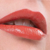 Close-up of lips wearing Benecos Natural Lipsticks 4.5g in a glossy, hydrating coral shade, slightly parted to reveal teeth. The background and area around the lips are a soft coral hue. Text below reads Soft Coral.