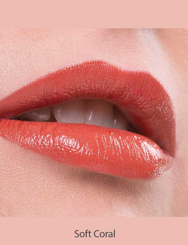 Close-up of lips wearing Benecos Natural Lipsticks 4.5g in a glossy, hydrating coral shade, slightly parted to reveal teeth. The background and area around the lips are a soft coral hue. Text below reads Soft Coral.