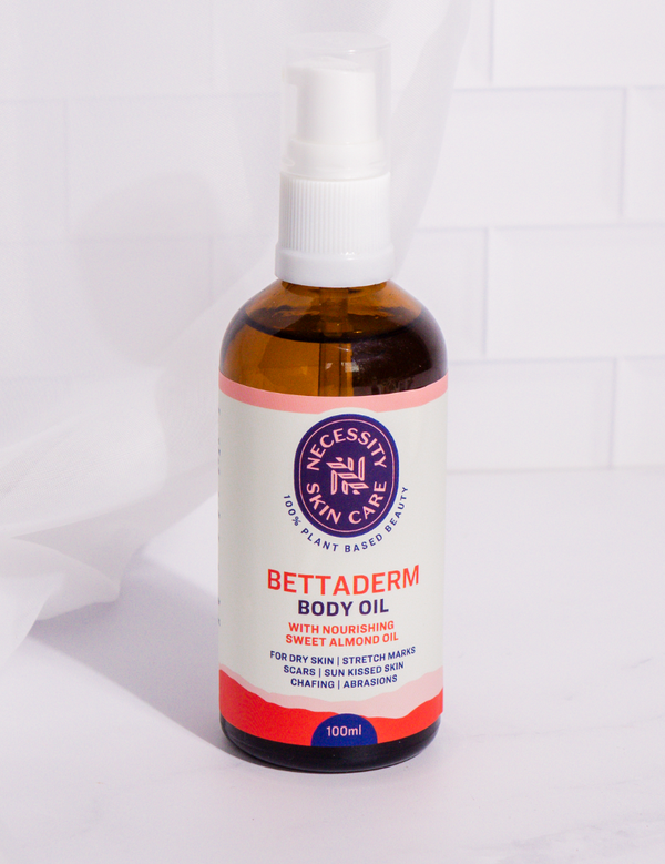 Amber Necessity Bettaderm Body Oil bottle with a white pump and pink-and-coral label, standing on a pale surface in front of white tiles and softly folded white fabric.