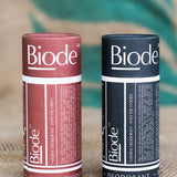 Two capped cylindrical Biode deodorant tubes standing side by side on coarse woven fabric: terracotta Into the Earth on the left and dark blue Into the Woods on the right.