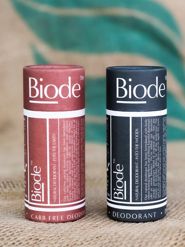Two capped cylindrical Biode deodorant tubes standing side by side on coarse woven fabric: terracotta Into the Earth on the left and dark blue Into the Woods on the right.