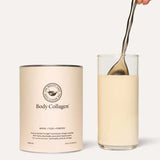 A hand stirs a creamy beige drink next to a The Beauty Chef Body Collagen™ Vanilla 260g container, showcasing the nourishing benefits of collagen peptides.