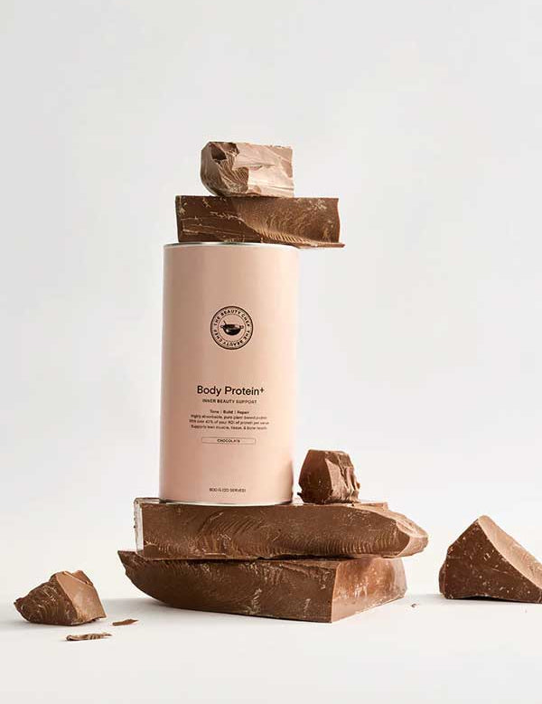 A cylindrical container of The Beauty Chef Body Protein+ Inner Beauty Support Chocolate 600g is surrounded and topped with large chocolate chunks, highlighting this plant-based protein powder against a plain light background.