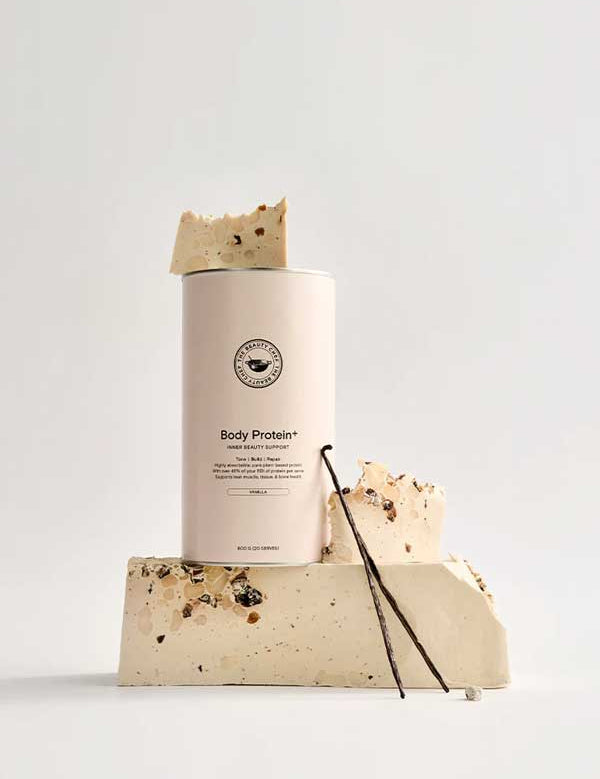 A beige canister of The Beauty Chef Body Protein+ Inner Beauty Support Vanilla 600g is surrounded by nougat-like protein bar chunks and vanilla beans, showcasing a plant-based protein powder with a complete amino acid profile on a light background.