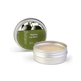 A silver metal tin of Botân Healing Lip Balm, with the lid leaning behind the open tin showing the beige balm inside. The lid features a green label with botanical illustrations.
