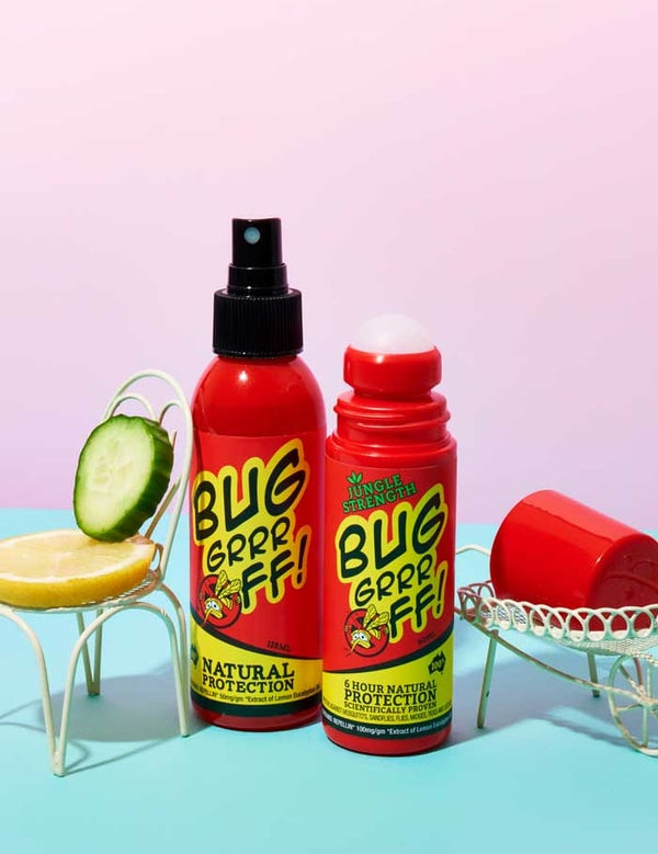 Two red bottles of Bug-Grrr Off 100% Natural Insect Repellent Jungle Strength Spray, with cucumber and lemon slices, sit on small white chairs against a pastel pink and blue background.