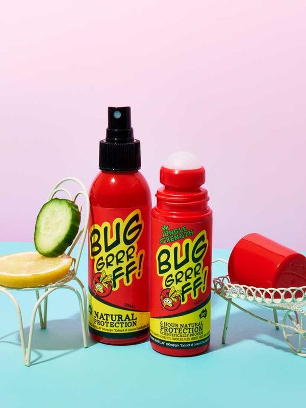 Two red bottles of Bug-Grrr Off 100% Natural Insect Repellent Jungle Strength Spray, with cucumber and lemon slices, sit on small white chairs against a pastel pink and blue background.