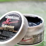 Open Elektra Charcoal Pomegranate Cleanser jar on a textured glass table, showing dark grey cream, with its black-labelled white lid leaning against it.