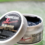 Open Elektra Charcoal Pomegranate Cleanser jar on a textured glass table, showing dark grey cream, with its black-labelled white lid leaning against it.