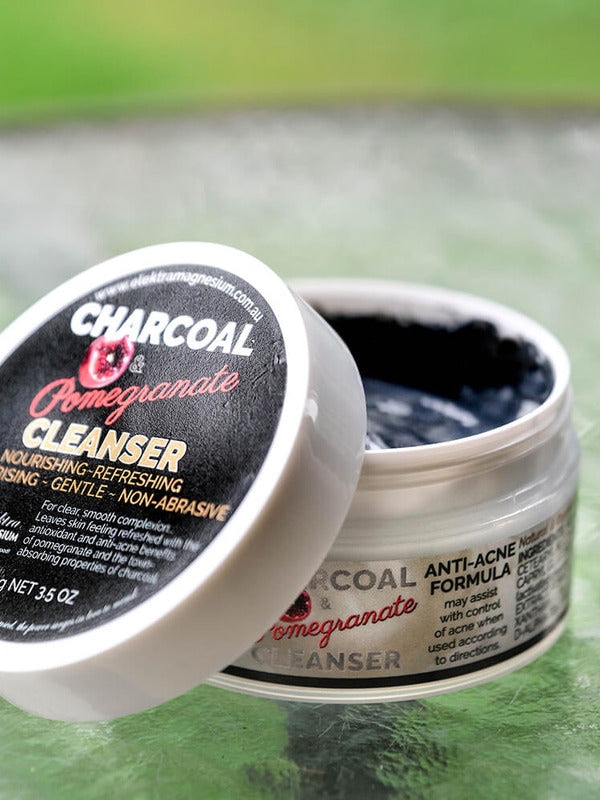 Open Elektra Charcoal Pomegranate Cleanser jar on a textured glass table, showing dark grey cream, with its black-labelled white lid leaning against it.