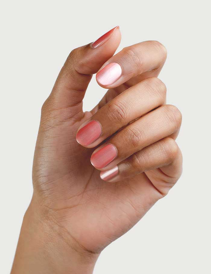 A hand with medium brown skin tone displays neatly manicured nails painted in Sienna Byron Bay Nail Polish - Charm 10ml, a glossy warm pink satin, against a plain light background.