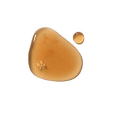 A glossy amber puddle of Dr Hauschka Clarifying Steam Bath liquid with a small separate round drop on white.