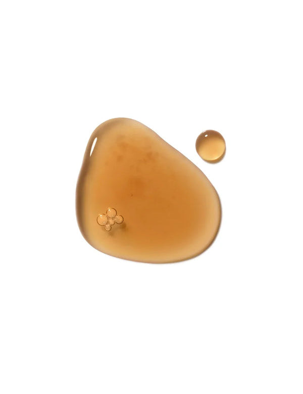 A glossy amber puddle of Dr Hauschka Clarifying Steam Bath liquid with a small separate round drop on white.