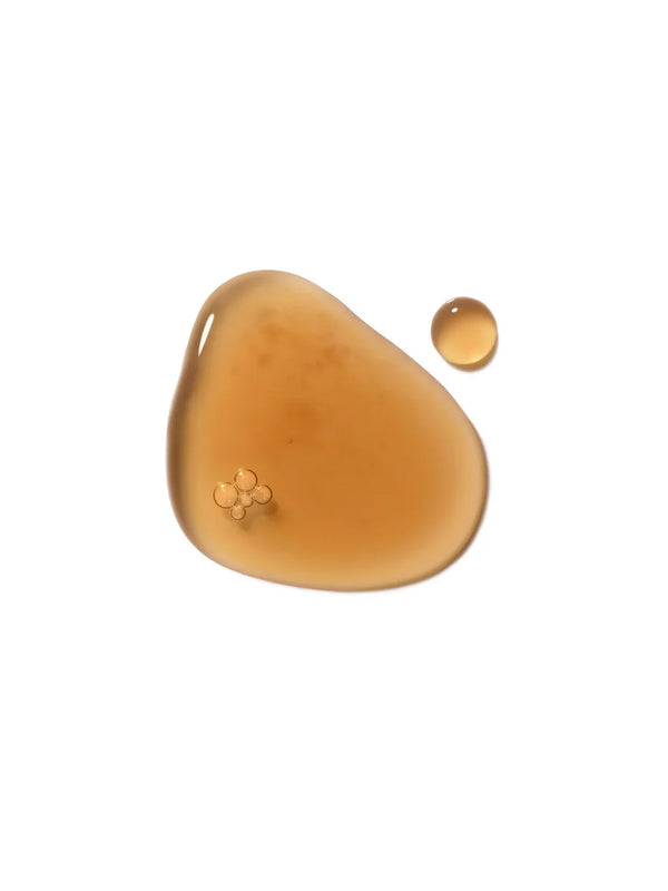 A glossy amber puddle of Dr Hauschka Clarifying Steam Bath liquid with a small separate round drop on white.