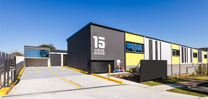 A modern commercial building with black, white, and yellow accents displays the address 15 Jubilee Avenue on its exterior. The driveway leads to a security gate under a clear blue sky.