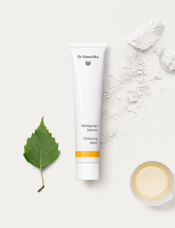 White Dr Hauschka Cleansing Balm tube arranged beside a green leaf, pieces of white chalky material with scattered powder, and a small dish of pale yellow liquid.