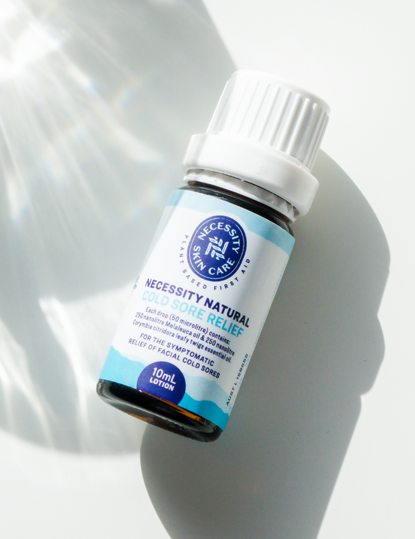 Small dark Necessity Natural Cold Sore Relief bottle lying diagonally on a pale surface, with a ribbed white cap, blue-bordered label and a long shadow beside rippling reflections of light.
