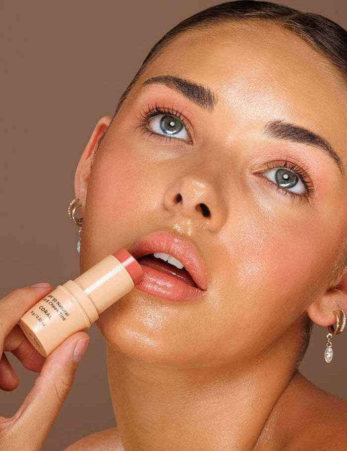 A blue-eyed woman with dewy skin and glossy lips poses against a brown background, holding Avocado Zinc SPF50 Natural Lip & Cheek Tint - Coral 9g near her lips. She has groomed brows and wears small hoop earrings.