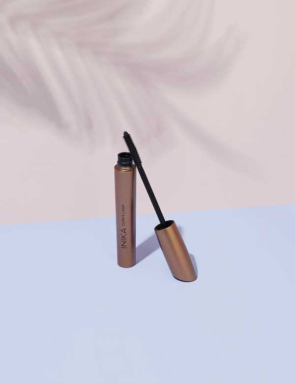 A tube of INIKA Organic Curvy Lash Mascara stands on a pastel blue surface with its black wand leaning against it, set against a light pink background—ideal for lifting and curling lashes all day.