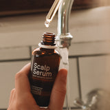 Hand holding an open amber Noosa Basics Scalp Serum bottle near a bathroom tap, with a drop forming at the tip of a glass pipette above it.