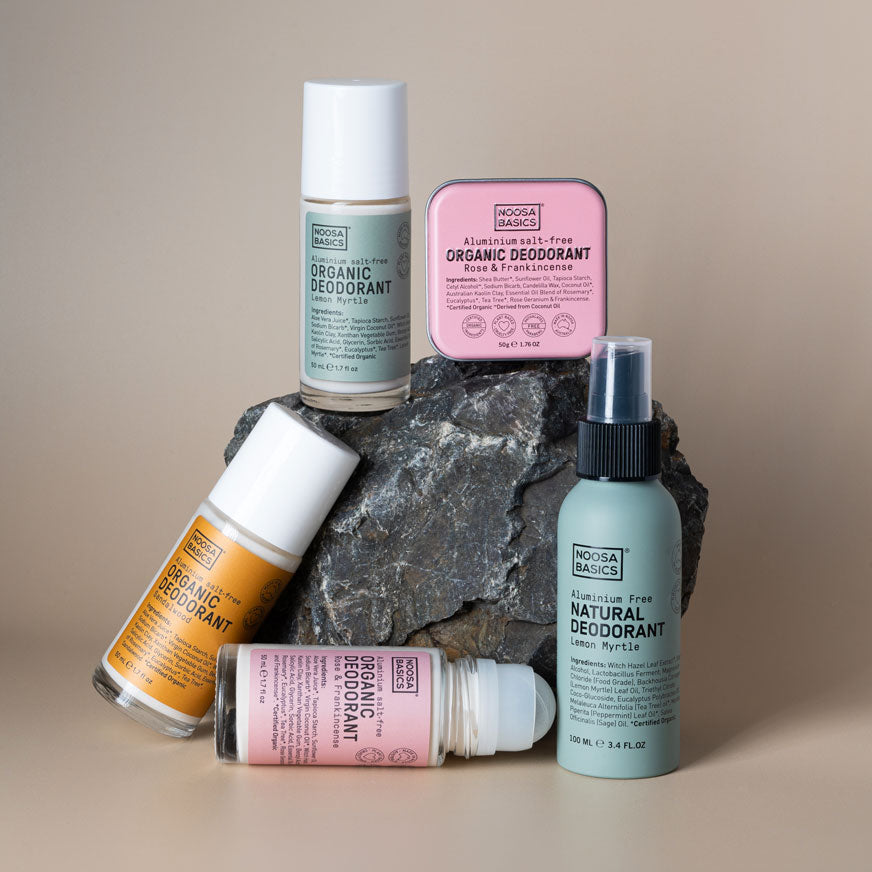 Five Noosa Basics deodorants arranged around a dark rock: three roll-on bottles with green, orange and pink labels, a square pink tin, and a pale green spray bottle.