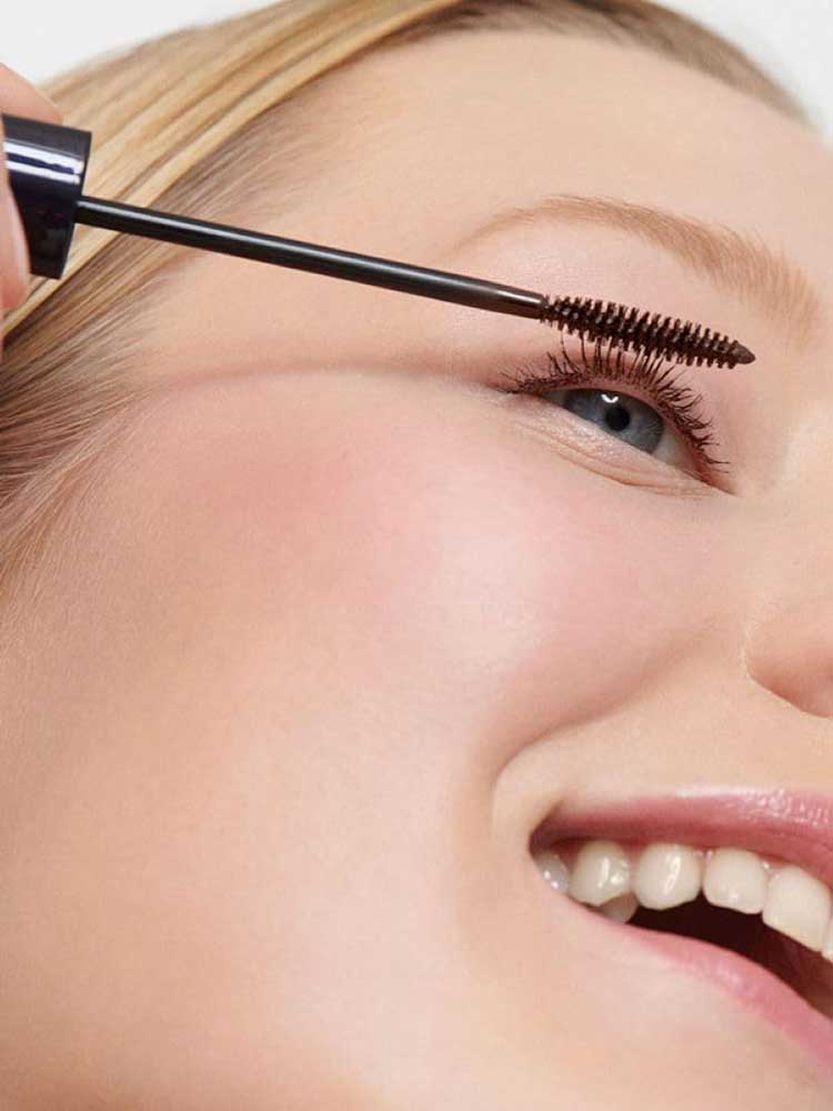 A close-up of a smiling person with blue eyes applying Dr Hauschka Defining Mascara 6ml—suitable for sensitive eyes—to their upper lashes. Their skin is smooth, and the mascara wand is held gently near their eyelashes.