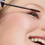 A close-up of a smiling person with blue eyes applying Dr Hauschka Defining Mascara 6ml—suitable for sensitive eyes—to their upper lashes. Their skin is smooth, and the mascara wand is held gently near their eyelashes.