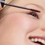 A close-up of a smiling person with blue eyes applying Dr Hauschka Defining Mascara 6ml—suitable for sensitive eyes—to their upper lashes. Their skin is smooth, and the mascara wand is held gently near their eyelashes.